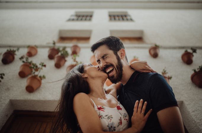 Joyful couple kissing and laughing while standing at street beside building
