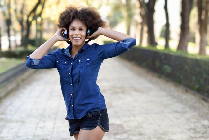 Mid shot of smiling female strolling outside in denim shirt while holding her headphones