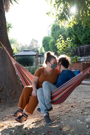 Romantic partners sitting in hammock with their faces close together wearing masks
