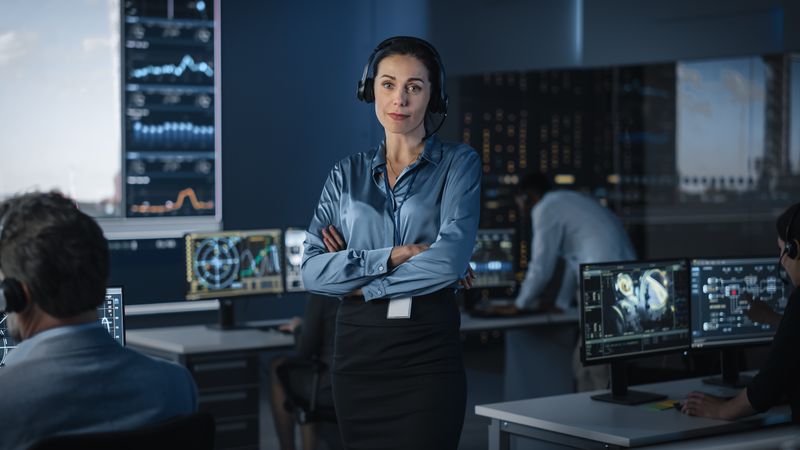 Confident Female Professional With Headset Supervises Team in High-tech Control Room.