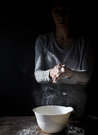 Woman clapping hands in flour near bowl and pack of eggs on table