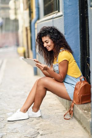 Smiling Arab woman looking at her digital tablet sitting outside a door, vertical