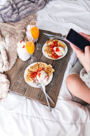 Woman with smartphone near appetizing waffles and tasty drinks on tray on bed