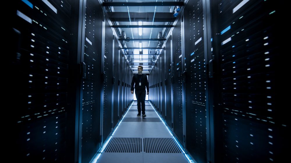 Shot of IT Engineer Walking Through Data Center Corridor with Rows of Rack Servers.