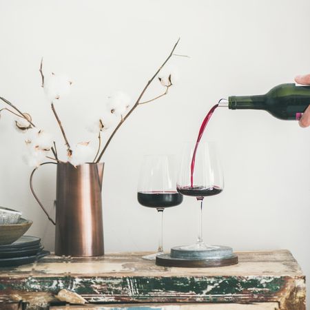 Wine glasses being poured from bottle with dried cotton in vase, square crop