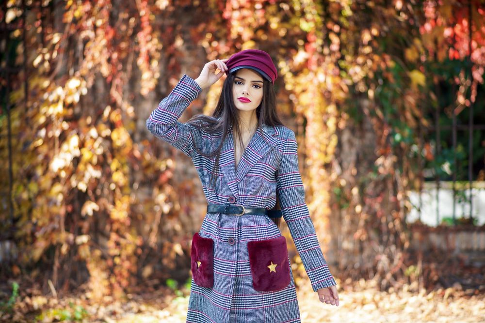 Female in warm winter clothes standing in park with colorful autumn leaves with hand to her hat