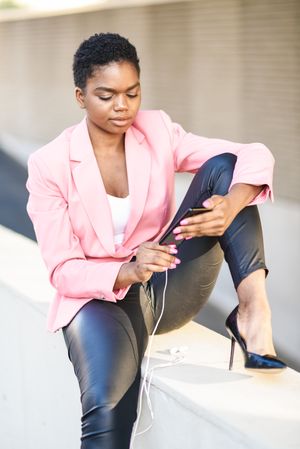 Female wearing suit with pink jacket sitting on ledge checking smartphone