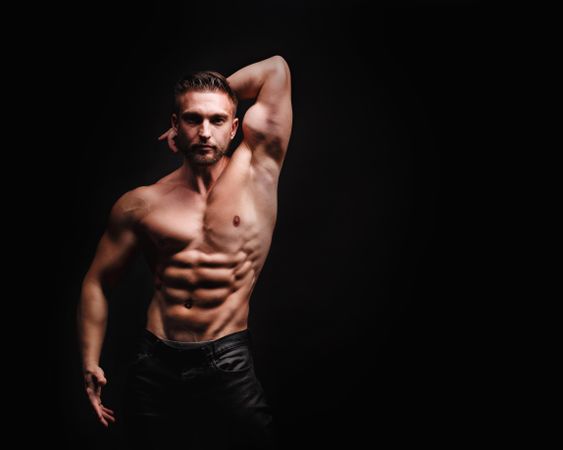 Bodybuilder competitor practicing arm and abs poses in dark studio, copy space