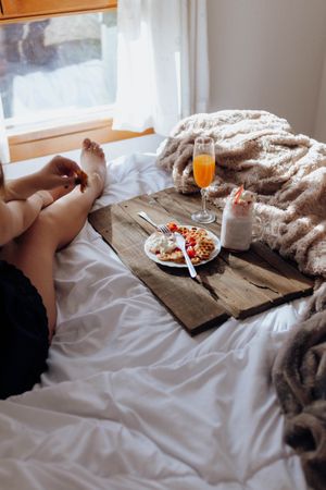 Young woman eating appetizing waffles and tasty drinks on tray on bed