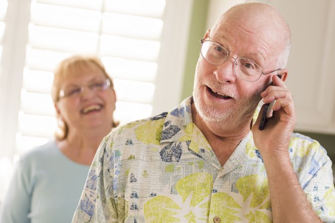 Older Adult Husband on Cell Phone with Wife Behind