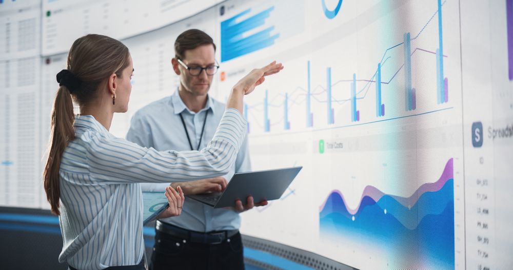 Male And Female Data Scientists With Laptop And Tablet Standing Next To Big Digital Screen With Graphs And Charts In Monitoring Office.