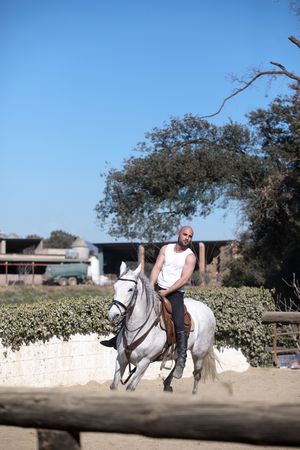 Man in sleeveless shirt riding horse on sandy ground around paddock