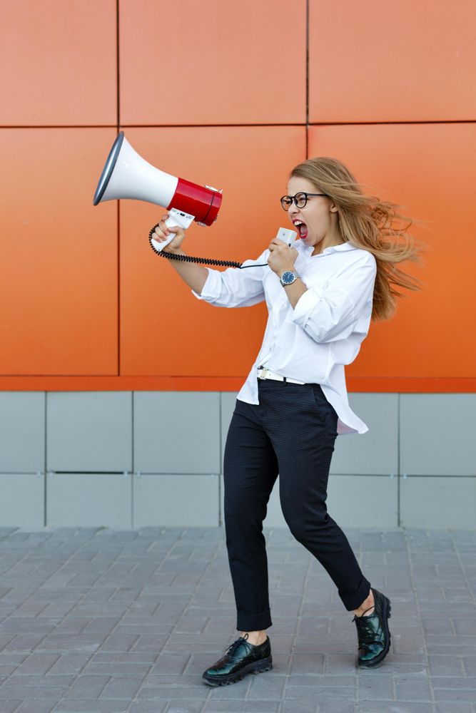 Woman in glasses in front of orange wall yelling into megaphone