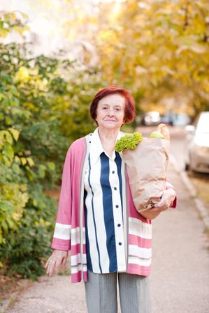 Older woman in pink cardigan holding grocery bag standing beside trees