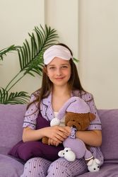 Girl seated on couch, holding teddy bear, sleep mask on.
