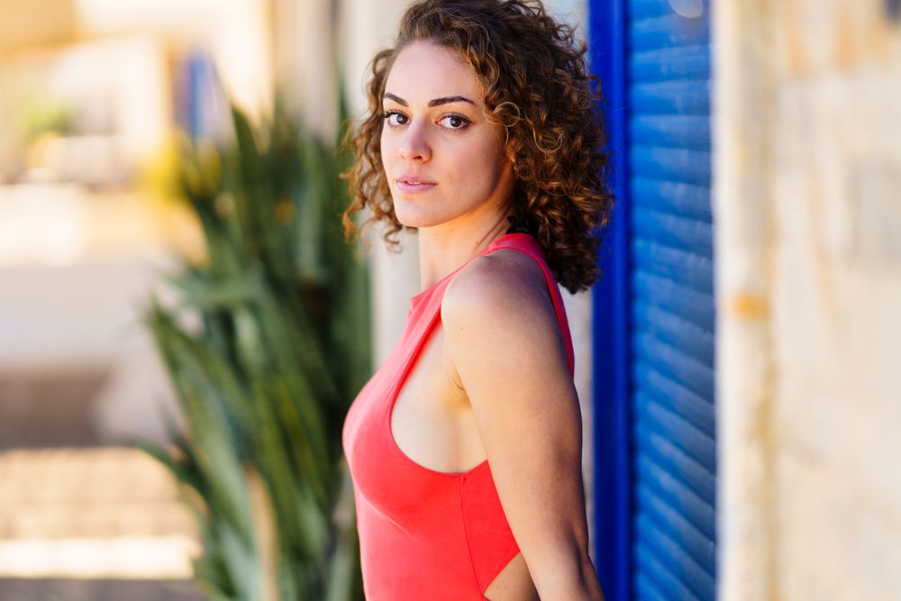 Side view of serious woman standing in front of blue shutters in red vest looking at camera
