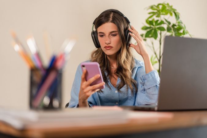 Young woman wearing headphones using smartphone while working on laptop at desk