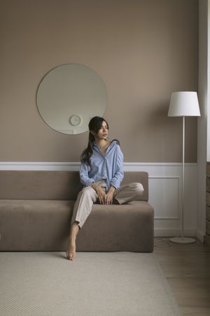 Young woman sitting at the end of a brown couch in a living room while looking out the window