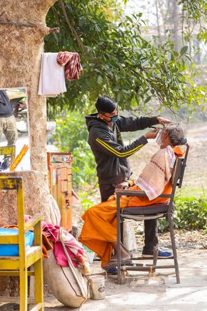 Barber with facemask shaving man's beard outdoor in India