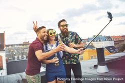 Three friends taking using selfie stick at goofy rooftop party bxQVX0