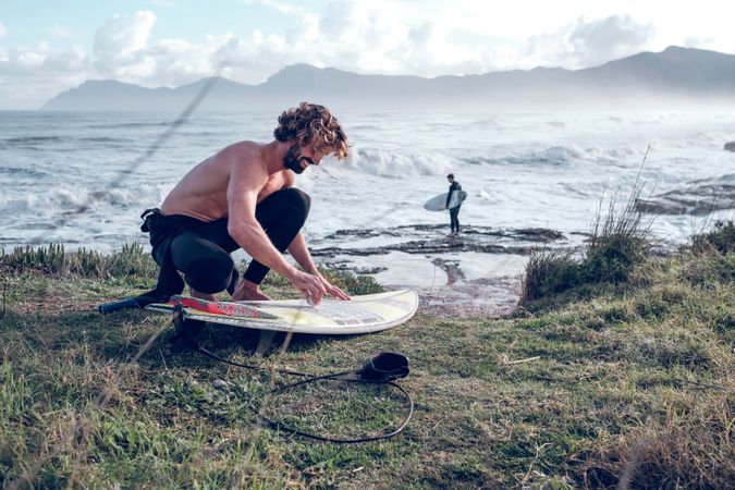 Surfer squatting on sea shore and covering surfboard with wax