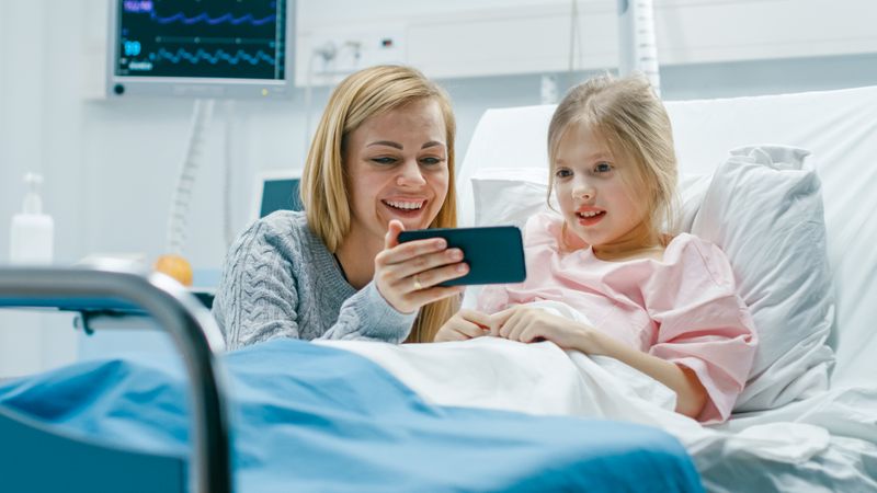 Cute Little Girl Lies on a Bed in the Children's Hospital, Her Mother Sits Beside, They Watch Cartoons/ Funny Videos on Smartphone.