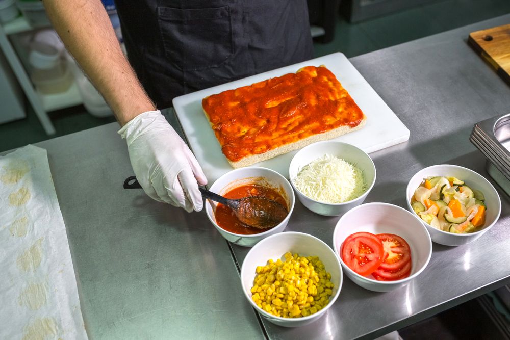 Unrecognizable male chef with gloves scooping tomato sauce to prepare pizza in restaurant kitchen