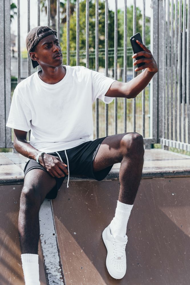 Sportive Black men taking selfie with smartphone while sitting on fence
