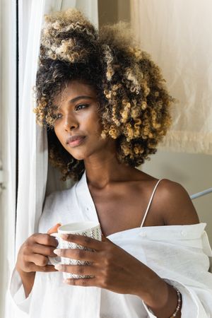Dreamy Black woman with cup of drink near window
