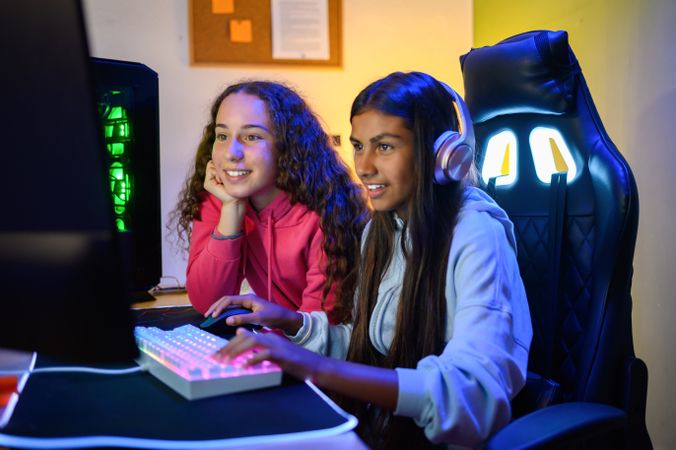 Cheerful teenage diverse sitting at table with gaming PC and playing game