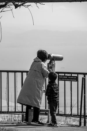 Grayscale photo of mother looking through binocular while son standing beside