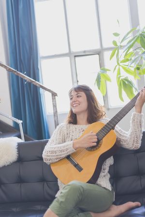 Happy female strumming guitar in living room of bright loft
