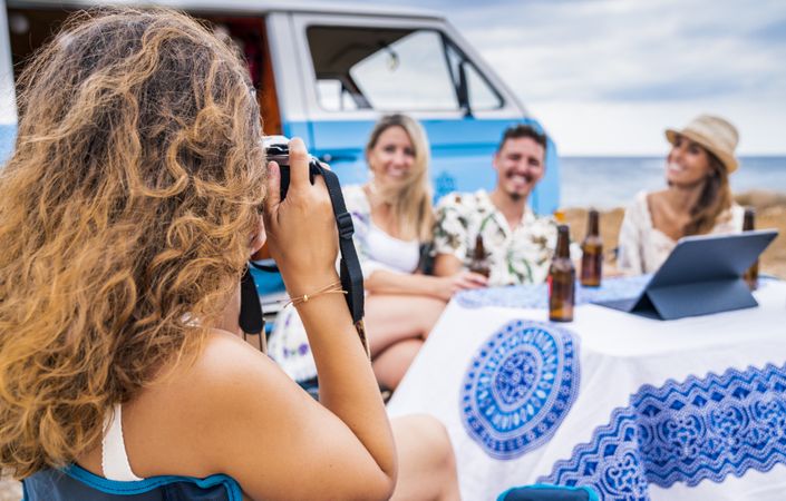 Joyful company sitting at table with drinks on beach while friend taking photo