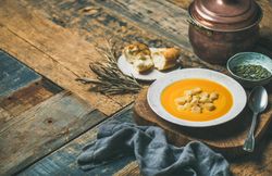 Pumpkin soup with croutons, bread, seeds, pot, spoon on wooden table with copy space