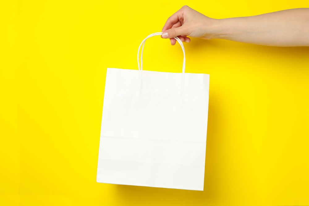 Female hand holding paper bag on yellow background, space for text