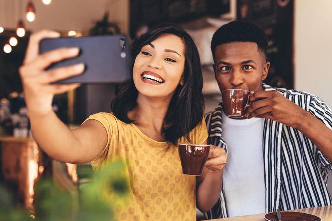 Woman taking selfie with friend making funny expressions while drinking coffee