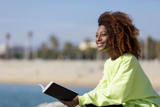 Happy female in bright green shirt relaxing on the seaside on sunny day with a book