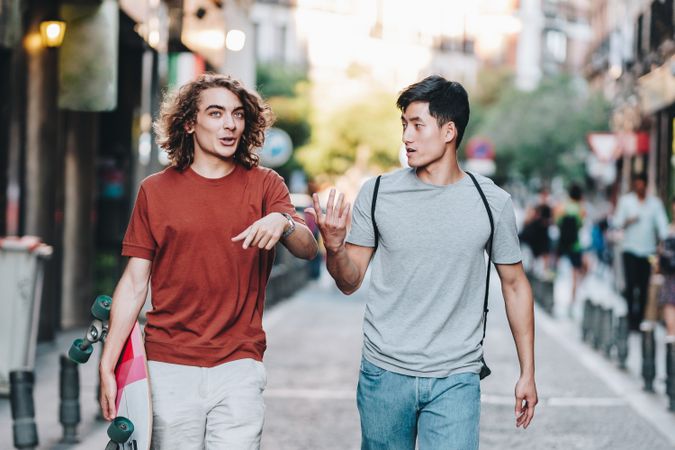 Cool multiracial male friends chatting while walking on street with longboard