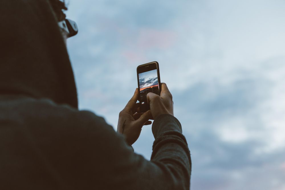 Rearshot of man on cliff taking photo on phone at sunset