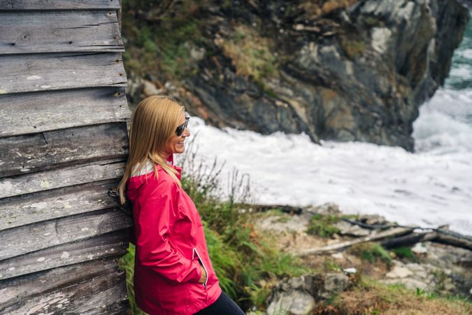 Woman in red raincoat leaning on house at shore looking at water
