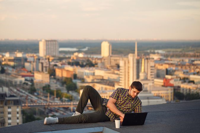 Man reclining on roof over looking skyline working on laptop with takeaway drink