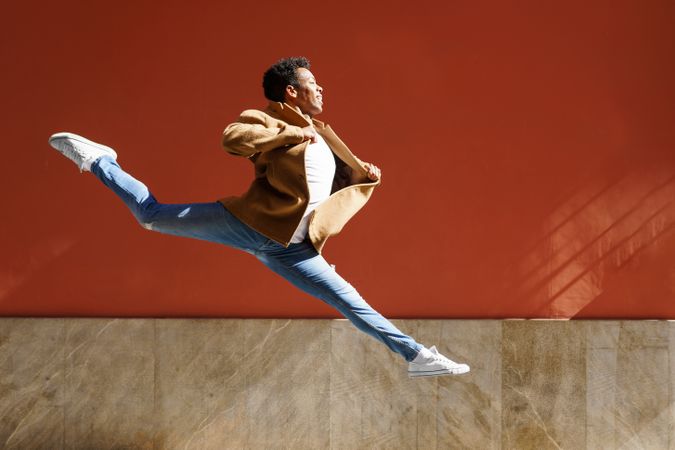 Graceful Black male in camel coat jumping against am outdoor wall