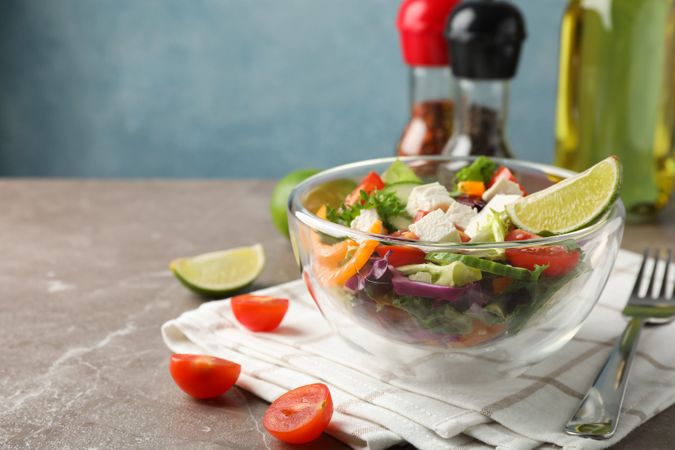 Glass bowl with salad, towel and spices on grey background, space for text