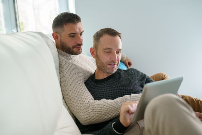 Two men relaxing together on sofa reading something on laptop