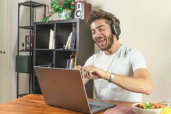 Young man laughing out loud while using a laptop with headphones at home.