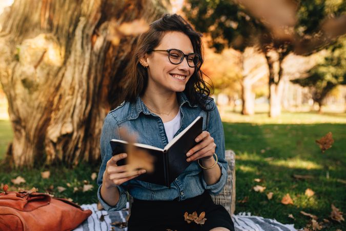 Smiling woman sitting at park holding a book with leaves falling around her