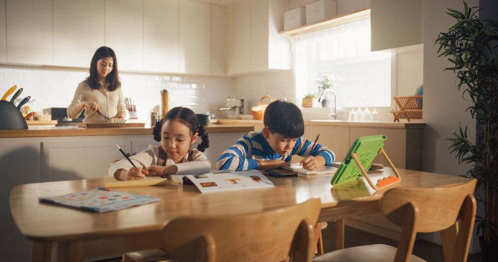 Portrait of a Little Korean Kids Sitting at a Kitchen Table at Home in the Morning While Their Mother is Cooking in the Background.