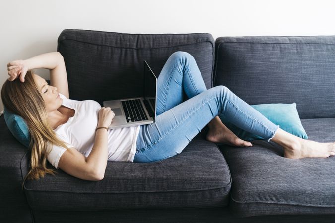 Pretty long haired female lying on sofa with laptop computer working remotely from home