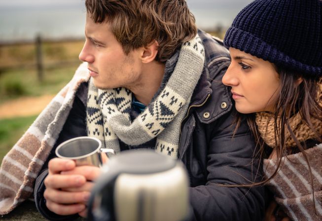 Couple wrapped in blanket on cold park bench with coffee flask sitting in silence