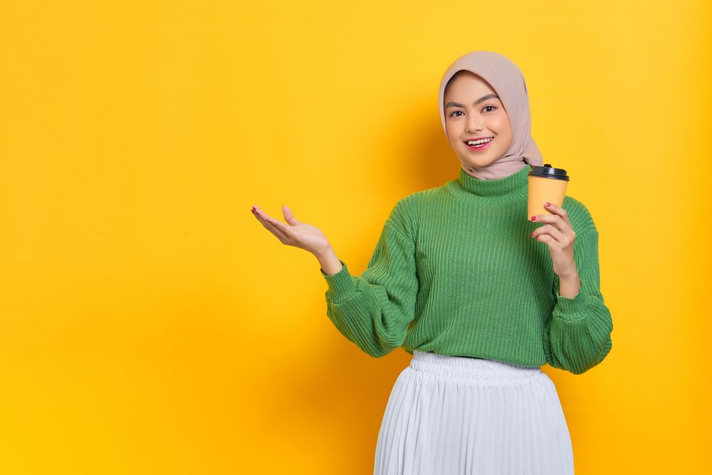 Woman in headscarf holding to go coffee cup and gesturing to the copy space